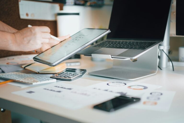 person using pen on ipad above a desk with a laptop, calculator, and printed data sheets